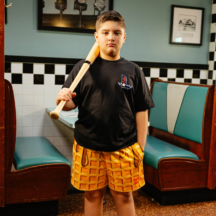 Person holding a baseball bat wearing waffle shorts and Black t-shirt in a diner setting