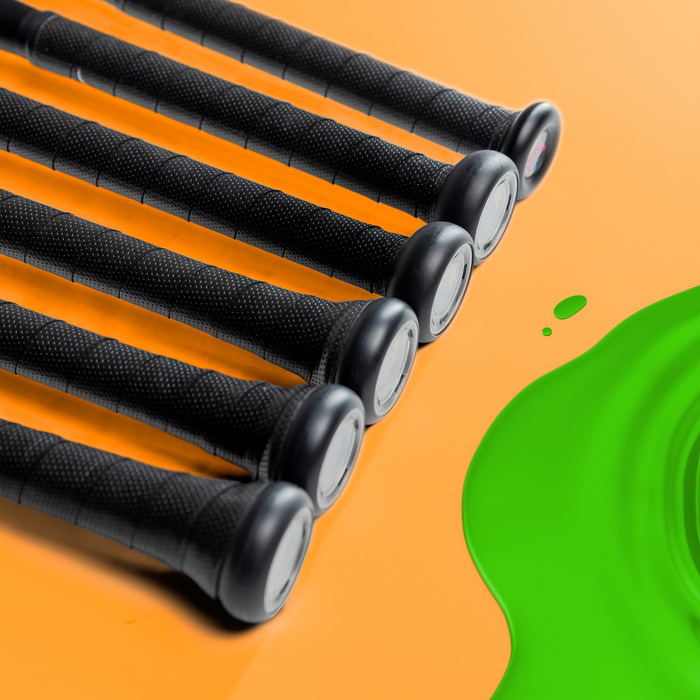 Six 2024 Victus Vibe (-5) USSSA Baseball Bats by Victus, featuring black one-piece aluminum handles, are aligned diagonally on an orange surface with a green paint splatter visible on the right.