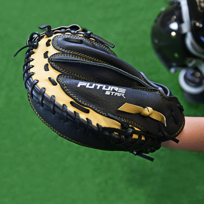 A close-up of the All Star Future Star 33.5" Baseball Catcher's Mitt (CM-FS-A) in black and tan durable leather, featuring FUTURE STAR branding, held over green turf with blurred baseball gear in the background.