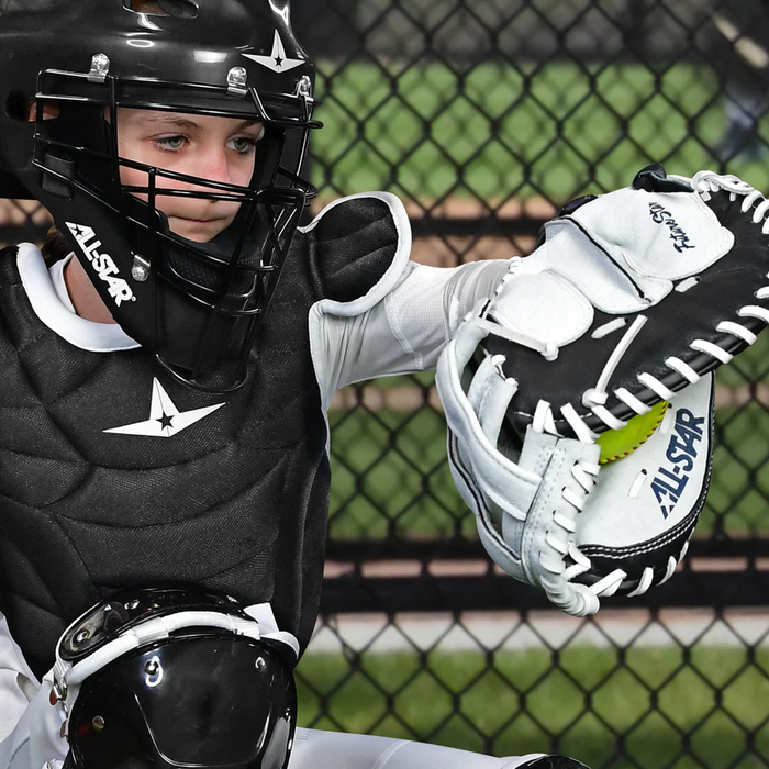 Wearing protective gear, a youth catcher focuses on catching a bright yellow softball with the All Star Future Star 34" Fastpitch Catcher's Mitt: CMW-FS-A, while a chain-link fence looms in the background during the game.