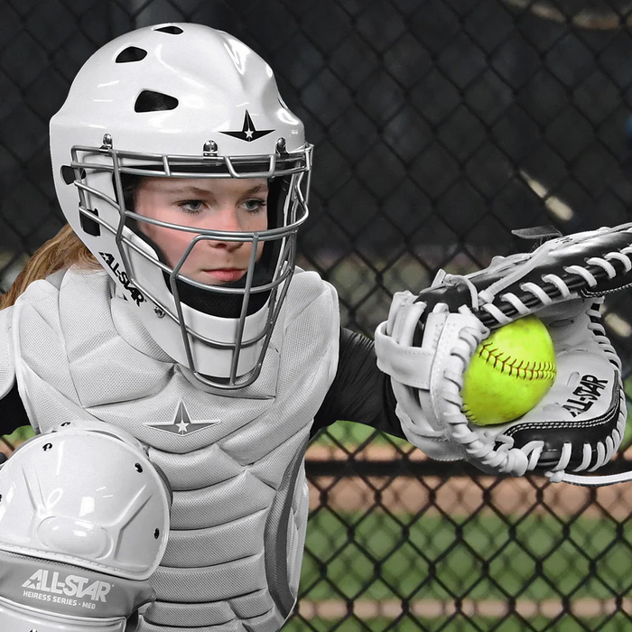 Wearing full white gear and a helmet, a travel ball catcher focuses as she catches a bright yellow softball with the All Star Heiress 34" Fastpitch Catcher's Mitt (CMW-H-A) by All Star against a chain-link fence on the field.