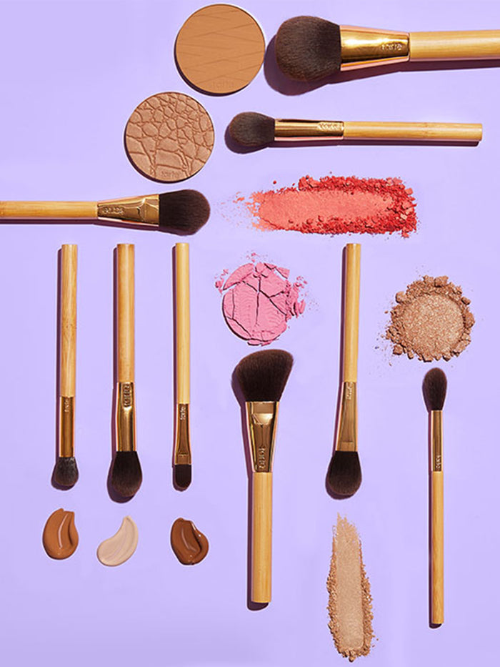 Makeup brushes with wooden handles are laid out with various cosmetic powders and creams, forming a neat arrangement on a lavender background. Text on the brushes: tarte.