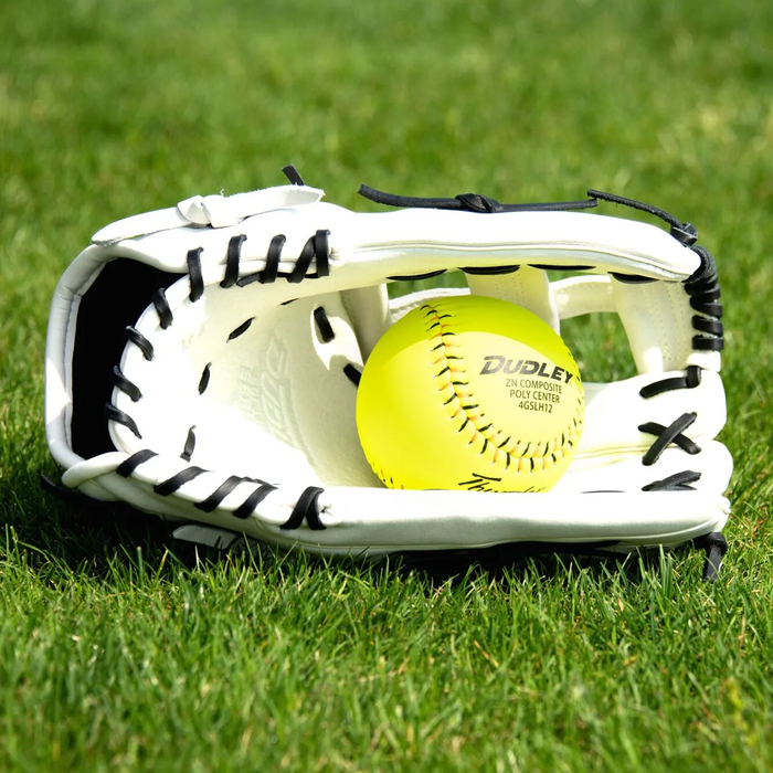 The Dudley 14" Slowpitch Glove: DFG14H, a white leather glove with black laces, lies open on green grass holding a bright yellow Dudley softball—ideal for slowpitch fielding.
