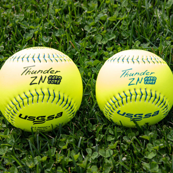 Two Dudley USSSA Thunder ZN Hard Core Pro W 11" 44/375 Composite Slowpitch Softballs (4U11H) rest on green grass, showing visible stitching and similar markings—ideal for slowpitch games.