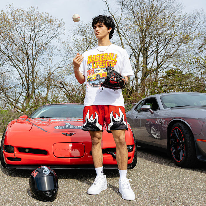 Young man tossing a baseball beside red Corvette and gray Dodge Challenger, wearing white BL101 race car tee and red flame mesh shorts, baseball glove in hand.