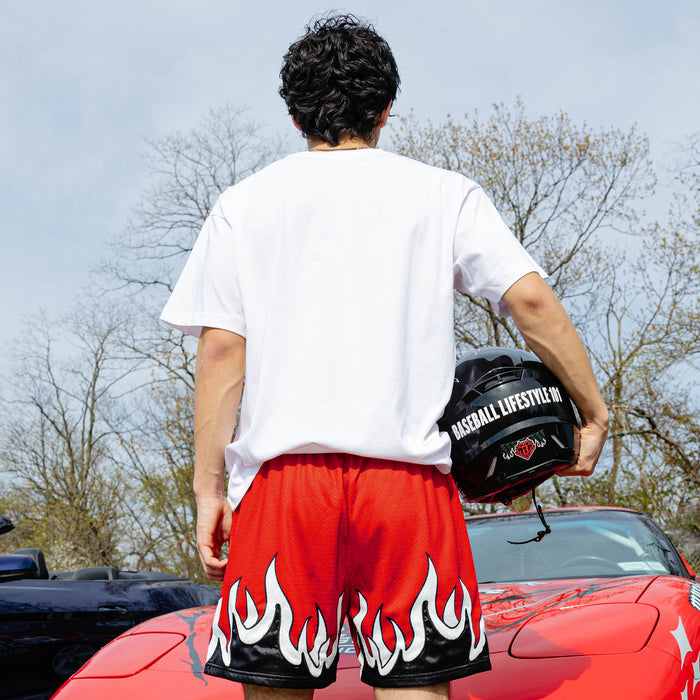 Back view of red flame mesh shorts with white and black flame hem graphic, model holding Baseball Lifestyle 101 helmet beside red Corvette.