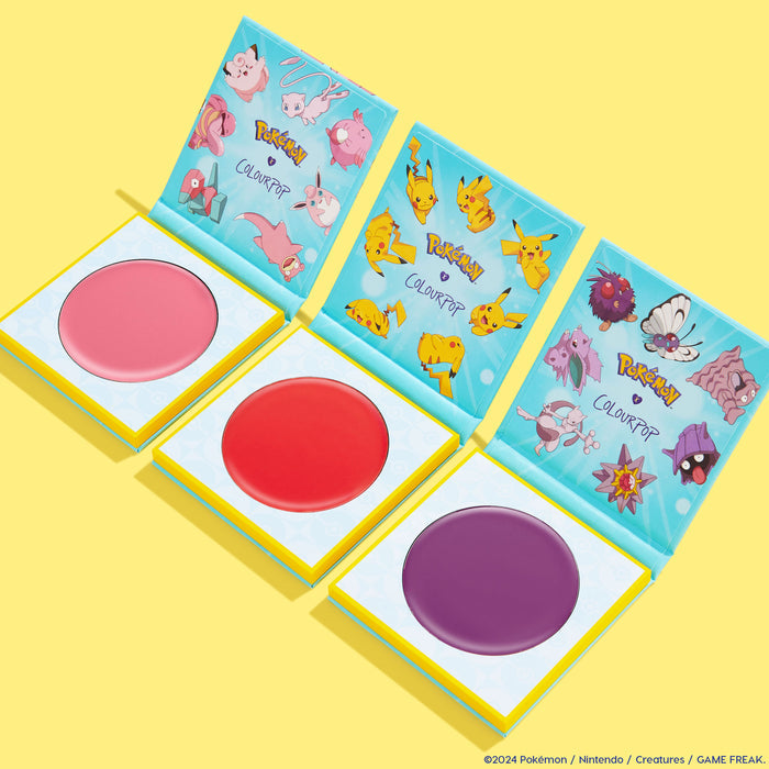 ColourPop Cream Blush Hypnosis compact opens to a deep purple pan, showcasing Pokémon collab lid art, set beside pink and red blush compacts on a bright yellow background for a playful, collectible cream cheek color.