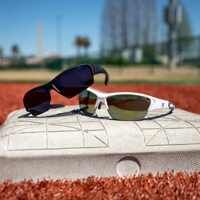 Two pairs of Marucci MV108 2.0 Performance Sunglasses (MSNV1082), one black and one white, sit atop a baseball base on a red dirt field with a sunlit, blurred outdoor background.