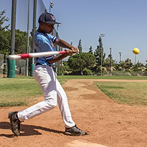 A young player in a blue uniform trains with PowerNet 2.8" Weighted Hitting and Batting Training Balls (6 Pack), swinging the bat and hitting a yellow ball on a sunny day at an outdoor baseball field.