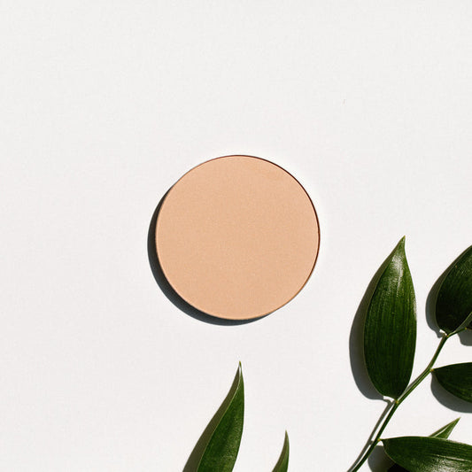 A pan of translucent pressed finishing powder placed on a white surface with leaves surrounding it.