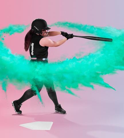 Wearing a Rip It Vision Pro Matte Fastpitch Softball Batting Helmet with Mask by Rip It, a softball player in black swings, sending green powder swirling near home plate against a pink and purple gradient background.