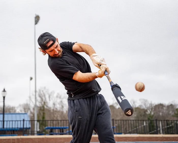 A man in athletic wear and a backward cap swings a baseball bat fitted with the Varo RAP Weighted Hittable Training Sleeve by Varo, making contact with a pitch outdoors on a cloudy day, with trees, a lamppost, and fence in the background.