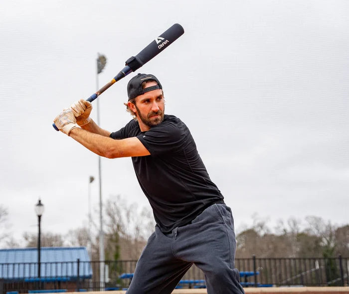 A man in a black shirt, gray sweatpants, and a backward cap readies his swing with the Varo RAP Weighted Hittable Training Sleeve by Varo on an outdoor field under cloudy skies, aiming for maximum swing speed. Trees and equipment are visible in the background.